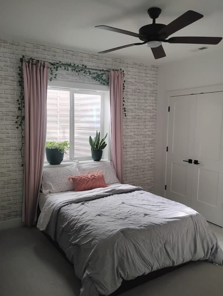 Cozy bedroom with white brick wall, pink curtains, grey bed, and plants on the windowsill.